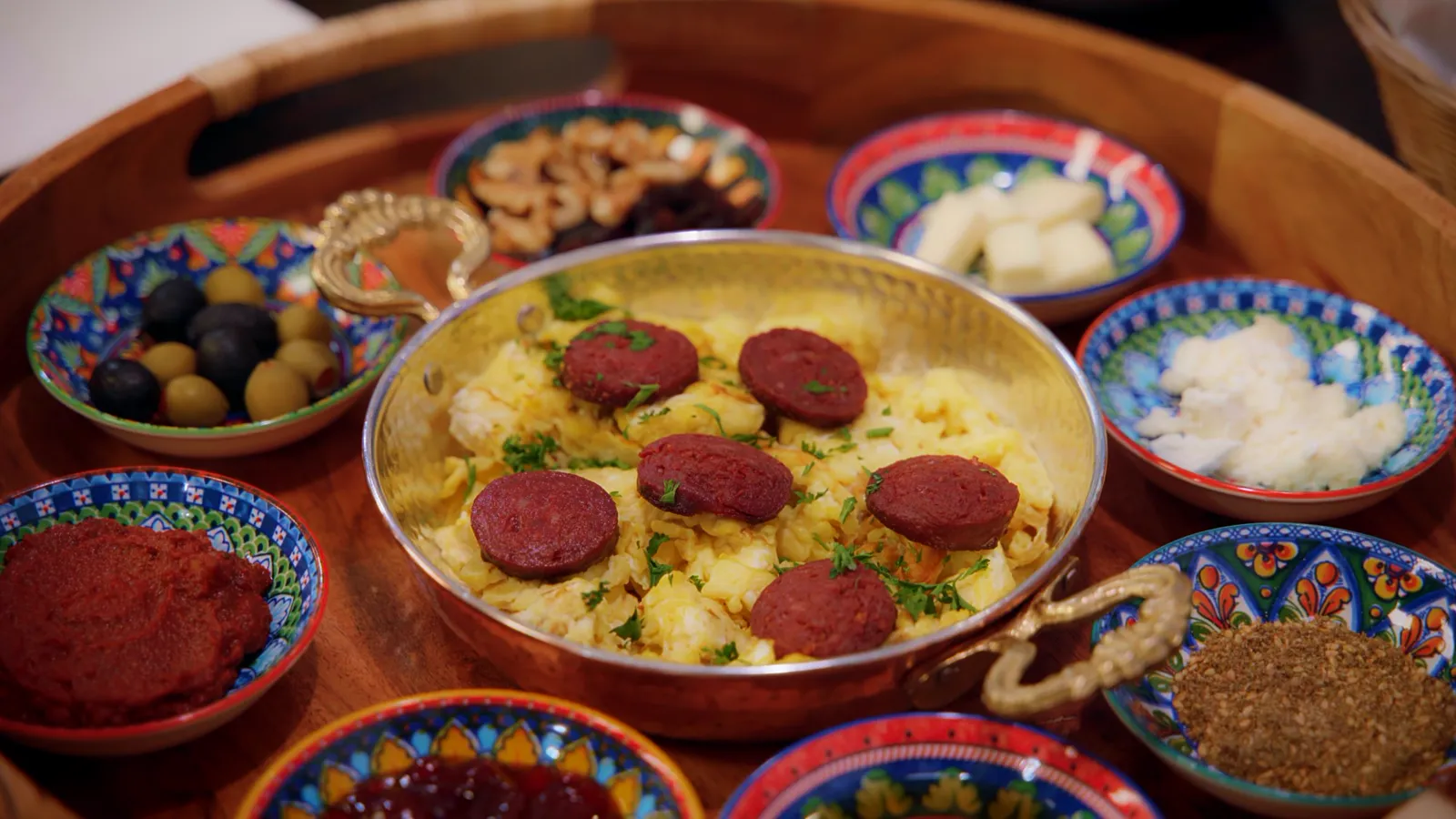 A Turkish breakfast spread with eggs and sausage in a copper pan surrounded by small bowls of olives, cheese, nuts, jam and za'atar