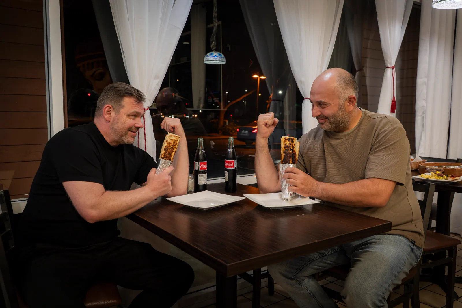 Two men at a restaurant table holding up toasted wraps side by side, flexing like they are comparing biceps